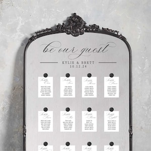 May include: A black framed mirror with a white background and black text that reads "be our guest" with the names "Kylie & Brett" and the date "10.12.24".  The mirror has eight white cards with black text that reads "Table One", "Table Two", "Table Three", "Table Four", "Table Five", "Table Six", "Table Seven", and "Table Eight" with a list of names under each table number.