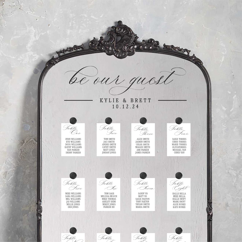 May include: A black framed mirror with a white background and black text that reads "be our guest" with the names "Kylie & Brett" and the date "10.12.24".  The mirror has eight white cards with black text that reads "Table One", "Table Two", "Table Three", "Table Four", "Table Five", "Table Six", "Table Seven", and "Table Eight" with a list of names under each table number.