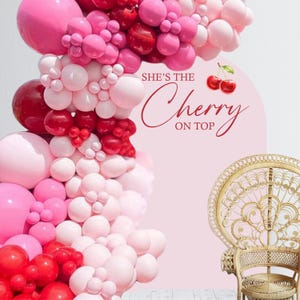 May include: A pink and red balloon arch with the text "She's the Cherry on Top" in red. A wicker chair is in the background.