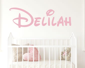 May include: Pink vinyl wall decal with the name "DELILAH" in a stylised font. The decal is above a white cot with a teddy bear and pillows.