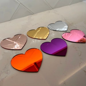 May include: Six heart-shaped decorative mirrors in various colors, including gold, pink, silver, purple, and orange. The mirrors are arranged on a white surface.