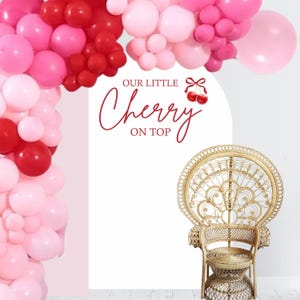 May include: A white backdrop with the text "Our Little Cherry On Top" in red lettering. A red bow with two red cherries is above the text. A pink and red balloon arch is above the text. A wicker peacock chair is in front of the backdrop.