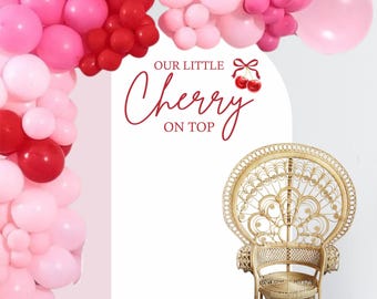 May include: A white backdrop with the text "Our Little Cherry On Top" in red lettering. A red bow with two red cherries is above the text. A pink and red balloon arch is above the text. A wicker peacock chair is in front of the backdrop.