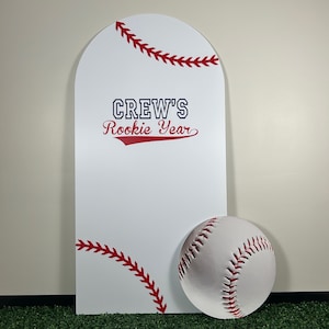 May include: White arch-shaped sign with red and white baseball stitching around the top. The sign says "CREW'S Rookie Year" in blue and red lettering. A white baseball with red stitching is in the bottom right corner of the sign.