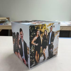 May include: A square photo block featuring family portraits on all sides. The images show a couple and a group of three people, all dressed in formal attire. The block is placed on a light-colored surface.
