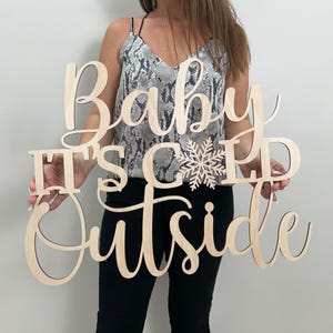 May include: A wooden sign with the phrase "Baby It's Cold Outside" in a cursive font. A snowflake design is between the words "Cold" and "Outside". The sign is made of light-colored wood.