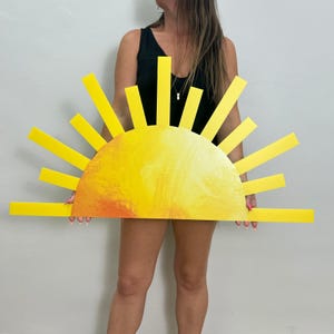 May include: A large, yellow sunburst decoration with a painted orange and yellow center and radiating yellow beams. The sunburst is held up against a plain white background.
