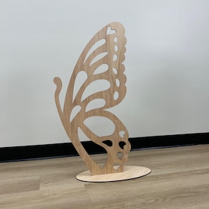 May include: A wooden butterfly cutout with intricate details, standing on a round base. The butterfly is light brown in color and has a delicate, whimsical design.