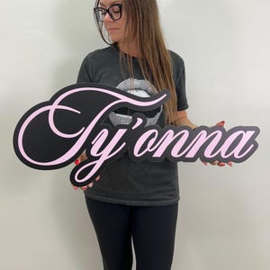 May include: A black sign with the name "Ty'onna" in a cursive font, outlined in pink. The sign is held by a person wearing a gray t-shirt and black pants.