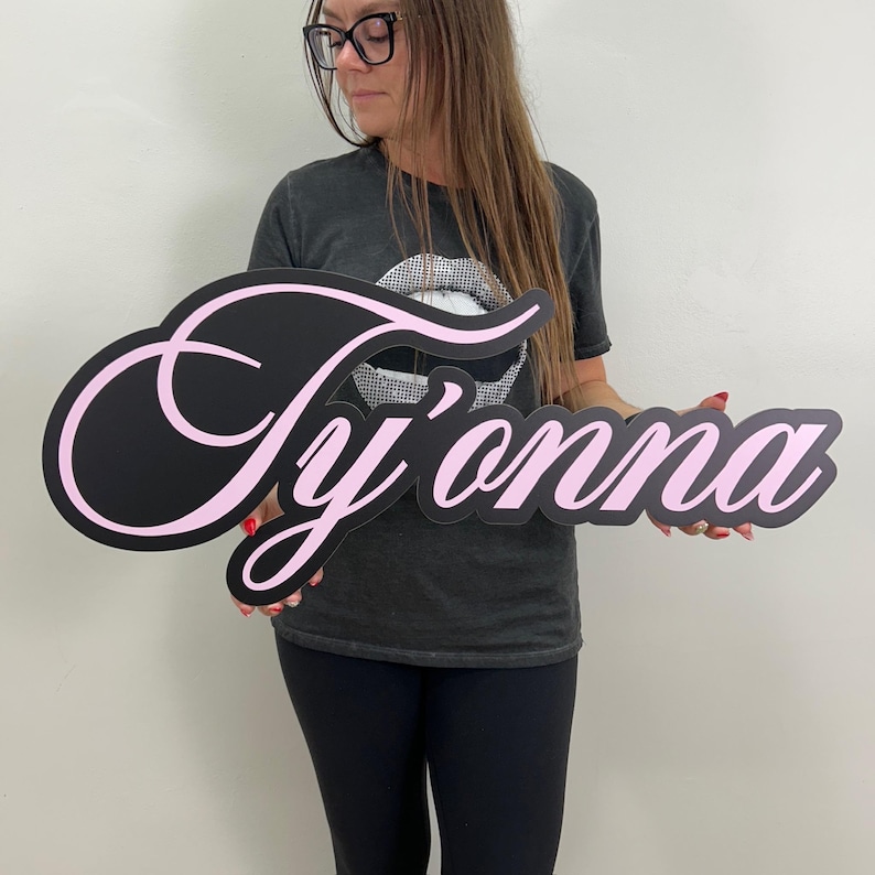 May include: A black sign with the name "Ty'onna" in a cursive font, outlined in pink. The sign is held by a person wearing a gray t-shirt and black pants.