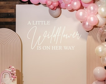 May include: A pink and white balloon arch with a sign that reads "A little wildflower is on her way". There is a cake with pink and white frosting and a rainbow on the bottom tier.