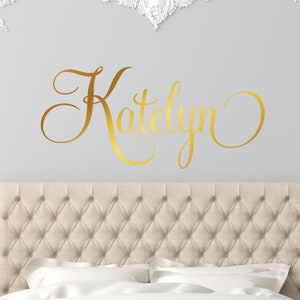 May include: Gold vinyl wall decal with the name "Katelyn" written in a cursive font. The decal is on a light gray wall above a beige headboard.