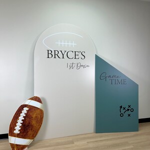 May include: A decorative football prop stands next to a white and teal backdrop. The backdrop features the text "BRYCE'S 1st Down" and "Game Time". A football-shaped prop is positioned on the left.