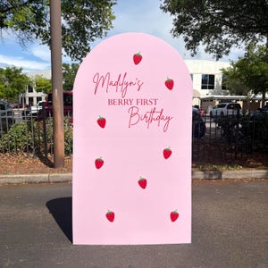 May include: A pink arch-shaped sign with the text "Madilyn's Berry First Birthday" in red script. The sign is decorated with red strawberry illustrations. The sign is outdoors.