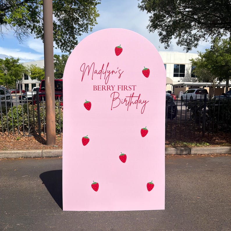 May include: A pink arch-shaped sign with the text "Madilyn's Berry First Birthday" in red script. The sign is decorated with red strawberry illustrations. The sign is outdoors.