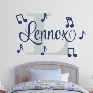 May include: A gray and white wall decal with the name "Lennox" in a cursive font and musical notes. The decal is above a wooden headboard with a blue and white patterned bedspread.