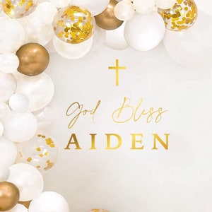 May include: White and gold balloon arch with a gold cross and the text "God Bless Aiden".
