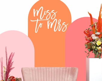 Puede incluir: Un sofá de terciopelo rosa se encuentra frente a un fondo con formas de arco en coral, rosa y blanco. Las palabras "Miss to Mrs" están escritas en escritura blanca. Arreglos florales en jarrones a ambos lados.