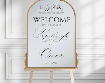 Puede incluir: Un cartel de bienvenida blanco con un marco dorado adornado, anunciando el baraat de Kayleigh y Omar el 08.11.2023. El cartel presenta la frase árabe "Te creamos en parejas" y su traducción al inglés.