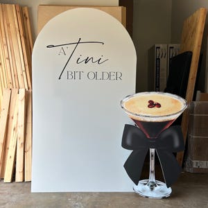 May include: A white arch-shaped sign with the text "A 'Tini Bit Older" in black script. A large cutout of a martini glass with a black bow is in front of the sign. The martini has a light brown foam and three coffee beans.