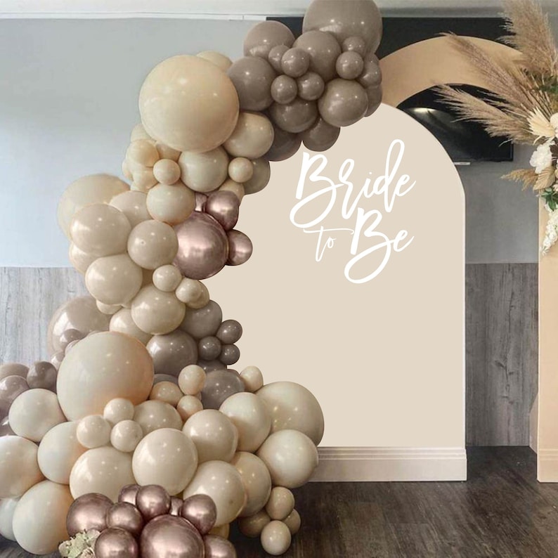 Pode incluir: Um arco de bal&otilde;es feito de bal&otilde;es bege, marrom e dourado rosa. O arco &eacute; decorado com as palavras "Bride to Be" em letras brancas.