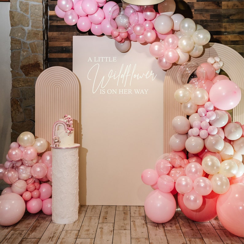 K&ouml;nnte beinhalten: Ein rosa und wei&szlig;er Kuchen mit einem Regenbogen obenauf steht auf einem wei&szlig;en Kuchenst&auml;nder. Der Kuchen steht vor einer hellrosa Wand mit dem Text "A little Wildflower is on her way". Die Wand ist mit rosa, wei&szlig;en und durchsichtigen Luftballons dekoriert.