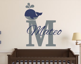 May include: A grey letter M with the name Mateo written in blue cursive above it. A blue whale with a grey tail and a water spout is sitting on top of the letter. The decal is on a white wall above a brown cot with a white and blue striped mattress.