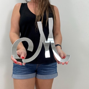 May include: A large, reflective silver letter "M" is held up in front of a white wall. The letter is in a cursive font and appears to be made of a shiny, metallic material. The letter is approximately 18 inches tall.