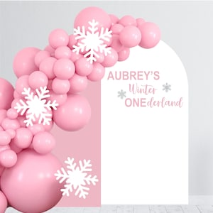 May include: Pink and white winter wonderland themed backdrop with a white arch and the text "Aubrey's Winter One-derland" with snowflakes. The backdrop is decorated with a garland of pink balloons.