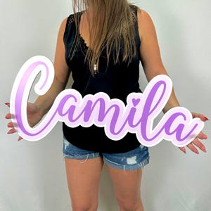 May include: A large, white, cursive name sign with a purple gradient. The name "Camila" is written in a decorative font with a small heart at the end of the name.