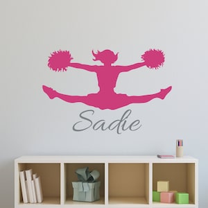 May include: Pink silhouette of a cheerleader with pom-poms, in a split pose, with the name Sadie written below.