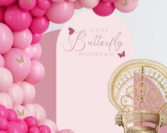 May include: A pink and white balloon arch with gold butterfly decorations. A white backdrop with the text "A Little Butterfly Is On Her Way" in pink script. A wicker peacock chair sits in front of the backdrop.