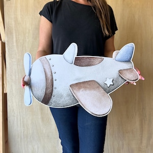 May include: A light gray and brown airplane cutout with a propeller, wings, and a star detail. The airplane has a vintage aesthetic, with a light blue propeller and tail accents. The cutout is held in front of a person.