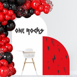 May include: A festive party scene with a balloon arch in red, black, and confetti-filled balloons. A white high chair with wooden legs is in front of a white arch with the text "ONE ROCKS". A red backdrop with black lightning bolt graphics completes the scene.