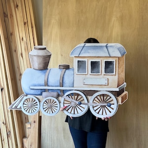 May include: A large, painted wooden train cutout in shades of blue, brown, and beige. The train features a detailed engine, smokestack, and wheels. The cutout is held up against a wooden background.
