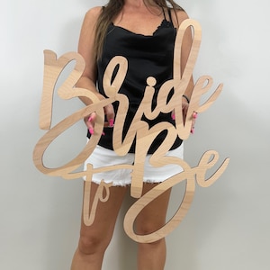 May include: A wooden sign in a cursive font that reads "Bride to Be". The sign is made of light-colored wood and is designed to be a decorative piece for a bridal event.