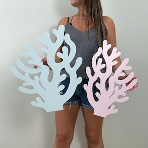 May include: Two coral-shaped wall decorations, one light blue and one pink. The decorative pieces have an organic, branching design, resembling coral formations. They are made of a solid material and are held up in front of a person.