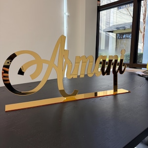 May include: A gold-colored, reflective acrylic sign that spells out the name "Armani" in a cursive font. The sign is mounted on a rectangular base, and the letters are connected. The sign is displayed on a dark surface.