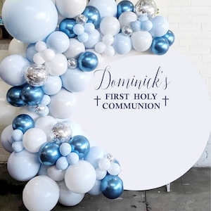May include: A balloon arch in shades of blue, white, and silver, with the text "Dominick's FIRST HOLY COMMUNION" in elegant script. The balloons are arranged in a semi-circle, creating a festive backdrop.
