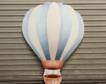 May include: A large, decorative hot air balloon cutout. The balloon features alternating vertical stripes of light blue and white. The basket is a reddish-brown color and is suspended by ropes. The background is a gray, corrugated metal surface.