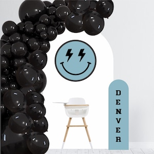 May include: Black and white balloon arch with a blue smiley face with lightning bolts for eyes. A white high chair with wooden legs is in the foreground. A blue sign with the word "DENVER" in black letters is on the right side of the image.