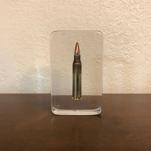 May include: A single bullet encased in a clear acrylic block. The bullet is a brass color with a copper tip.