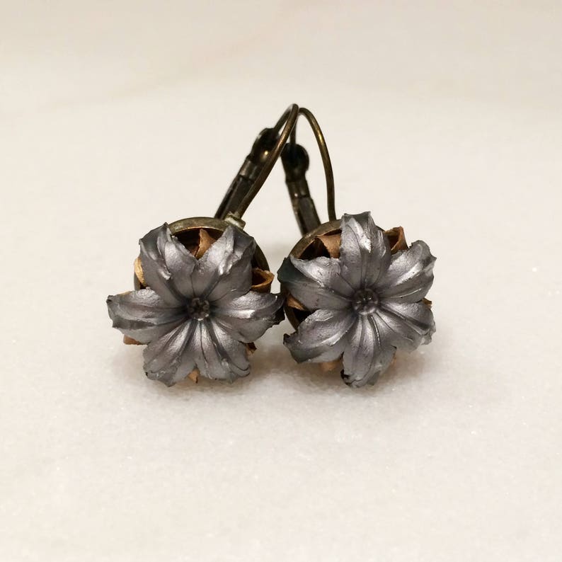 May include: A pair of silver bullet earrings with a flower design. The earrings are made from real bullet casings and have a bronze-colored hook.