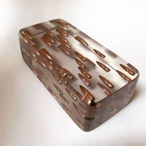 May include: A clear rectangular block of resin with numerous copper-colored bullet casings embedded inside. The casings are arranged in a random pattern, creating a unique and eye-catching design.