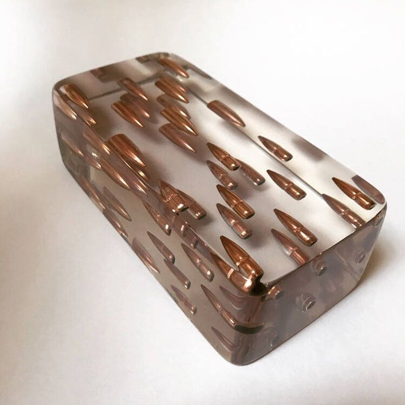 May include: A clear rectangular block of resin with numerous copper-colored bullet casings embedded inside. The casings are arranged in a random pattern, creating a unique and eye-catching design.