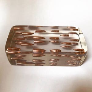 May include: A clear rectangular block filled with copper-colored bullets. The bullets are arranged in rows and columns, creating a geometric pattern.