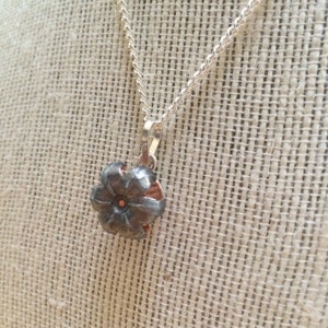 Può includere: Una collana con catena d'argento con un ciondolo realizzato da un bossolo di proiettile riutilizzato. Il bossolo di proiettile ha la forma di un fiore con un centro in rame.