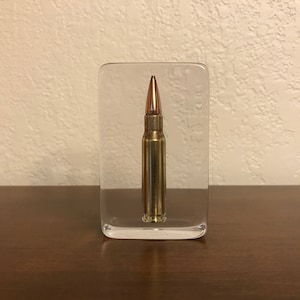 May include: A single bullet encased in a clear acrylic block. The bullet is gold and copper coloured.