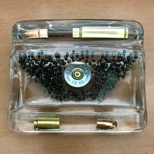 May include: A clear glass display case with a shotgun shell and two bullet casings. The shotgun shell is a Remington 12 GA. The case is filled with small black pellets.