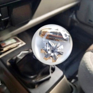 May include: Clear, spherical shift knob with a bullet and a silver and gold starburst design encased inside. The knob is mounted on a black gear shift lever in a vehicle's interior.